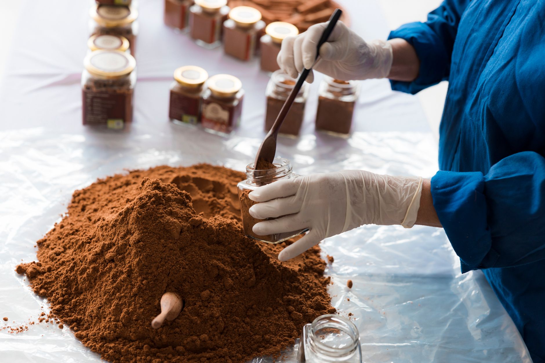 Hands wearing gloves packaging ground carob powder in glass jars with a wooden spoon.